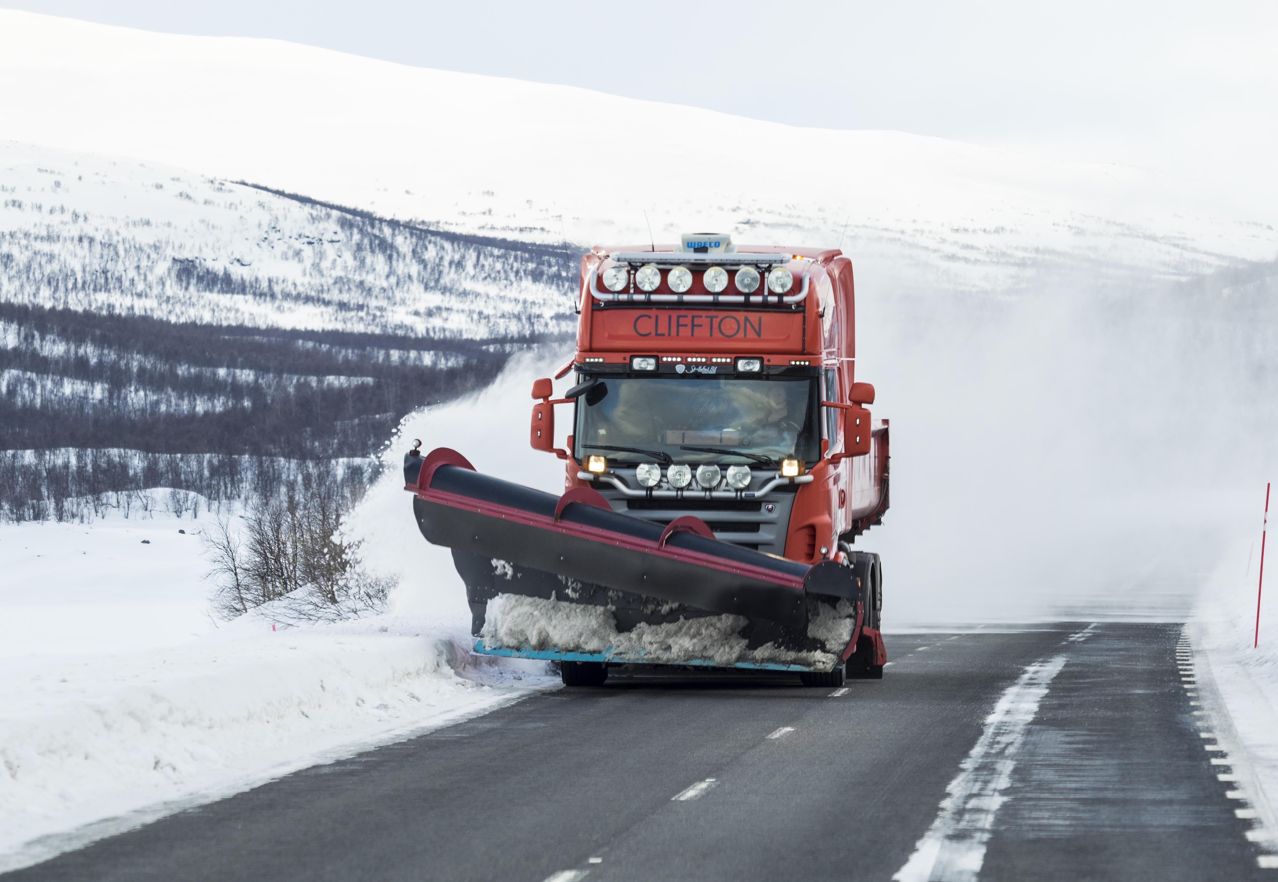 Bild på en plogbil som kör framåt på en asfaltsväg. I bakgrunden syns ett snötäckt berg och på sidan av vägen ligger ett metertjockt lager av snö.