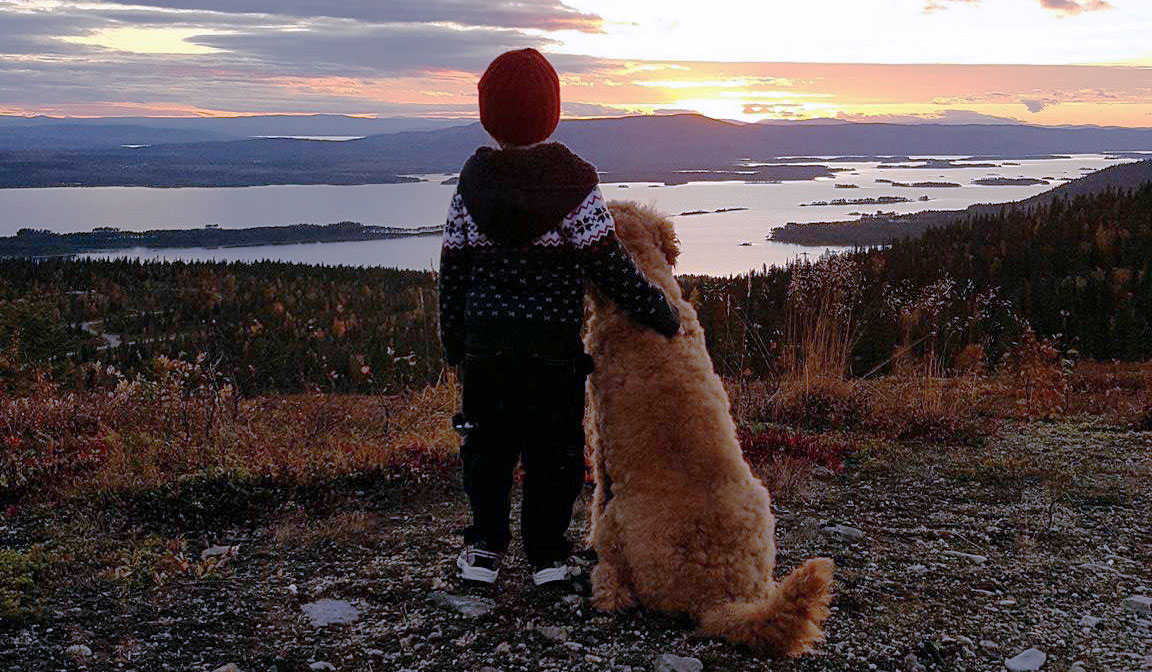 En ung pojke står med armen runt en hund på ett berg. De blickar ut över en sjö med fjäll i bakgrunden.