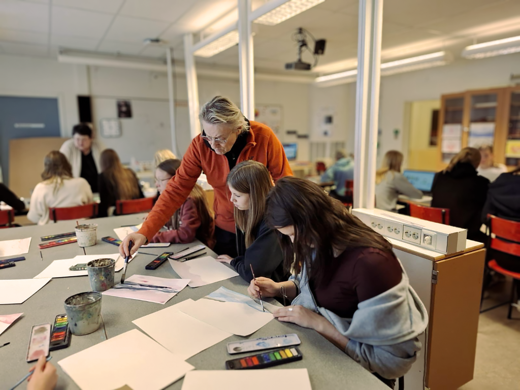 Lars Lerin står i ett klassrum, böjd över bordet med elever och instruerar dem i deras målande.