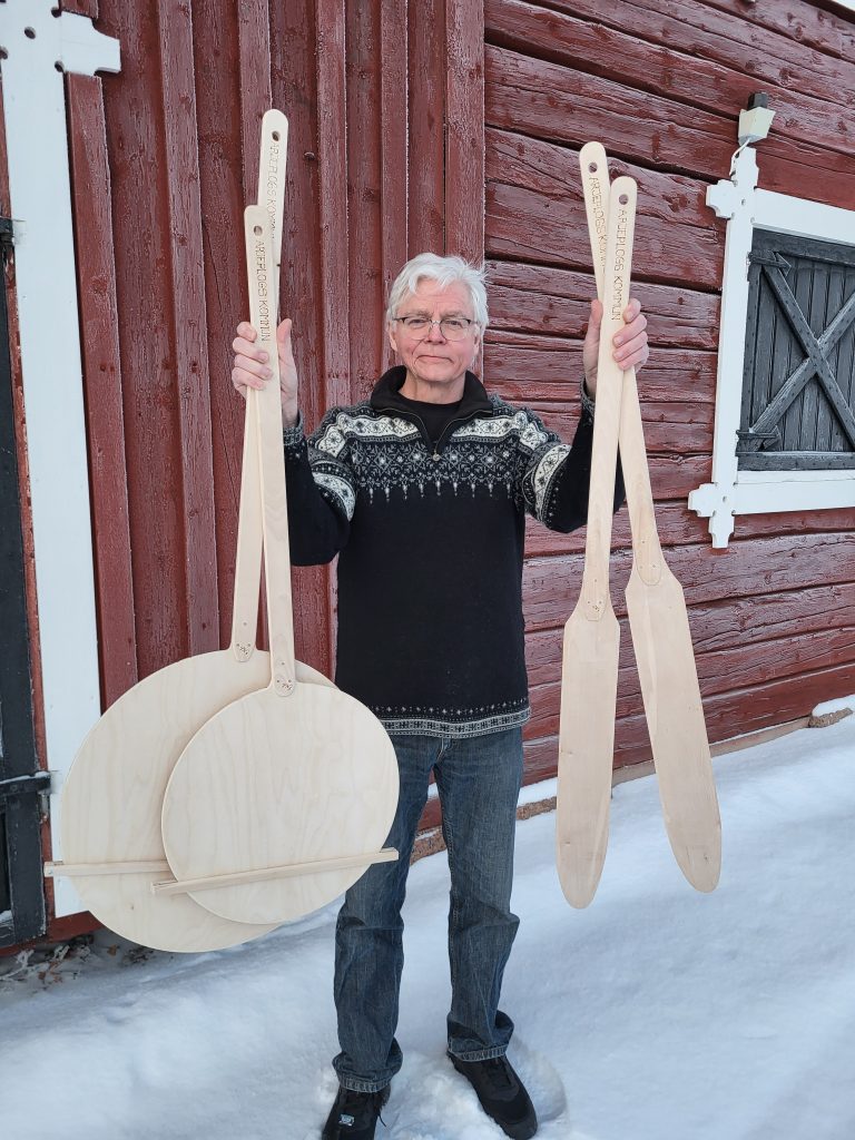 Peter Nilsson håller upp spjälkar och fjölar i björk, framför en faluröd husvägg, vintertid.