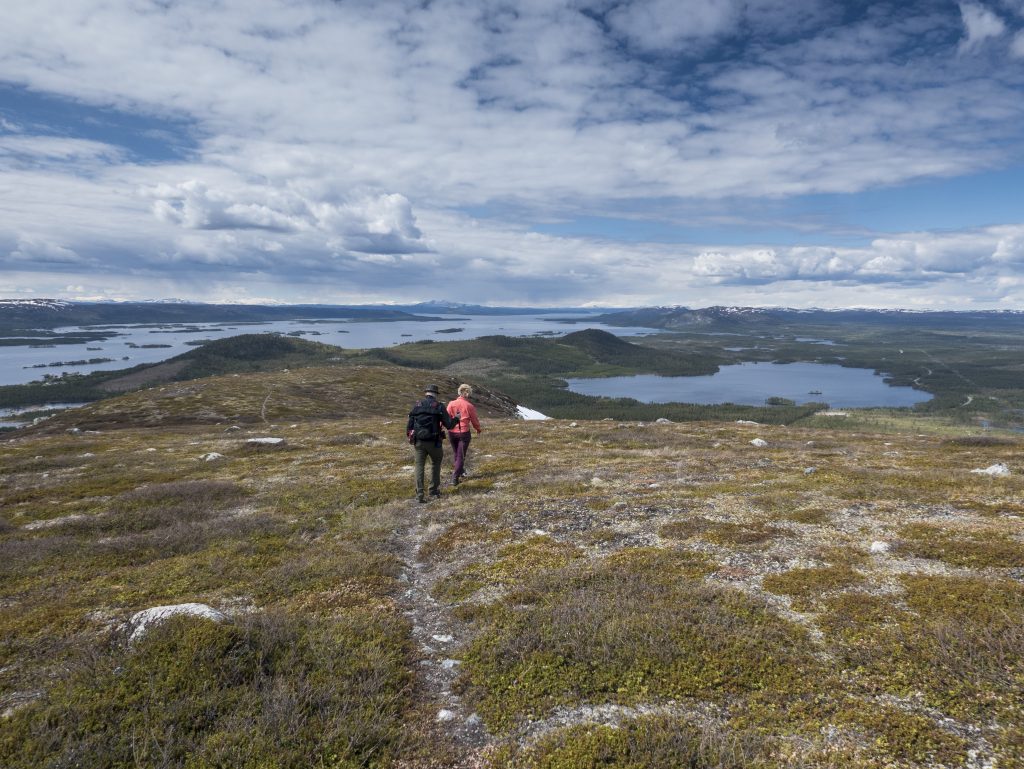 Två personer som vandrar längs en stig uppe på en fjälltopp. Sjöar i bakgrunden.