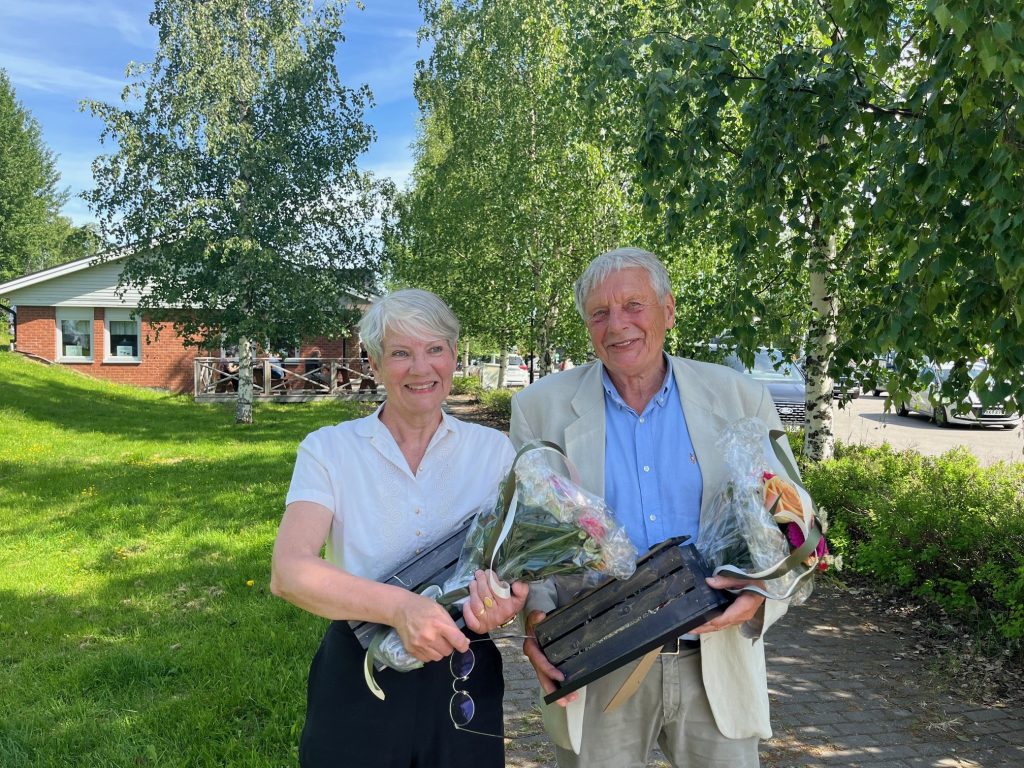 Ingela Bergman till vänster och Christer Andersson till höger, håller i varsin blomma, inslagen gåva och diplom, en vacker sommardag.