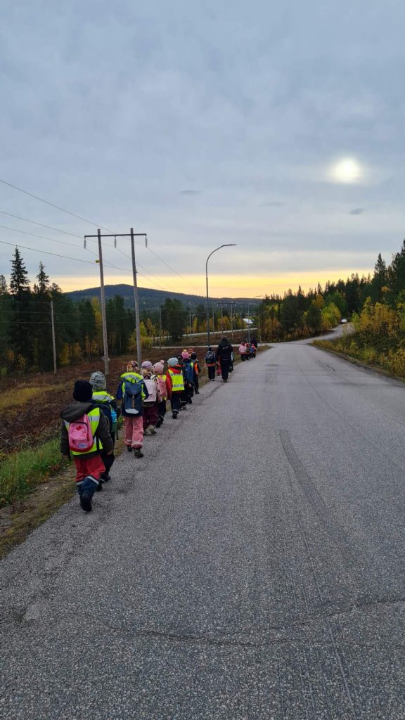 Förskolebarn på led i varselvästar i skymningen längs en väg.