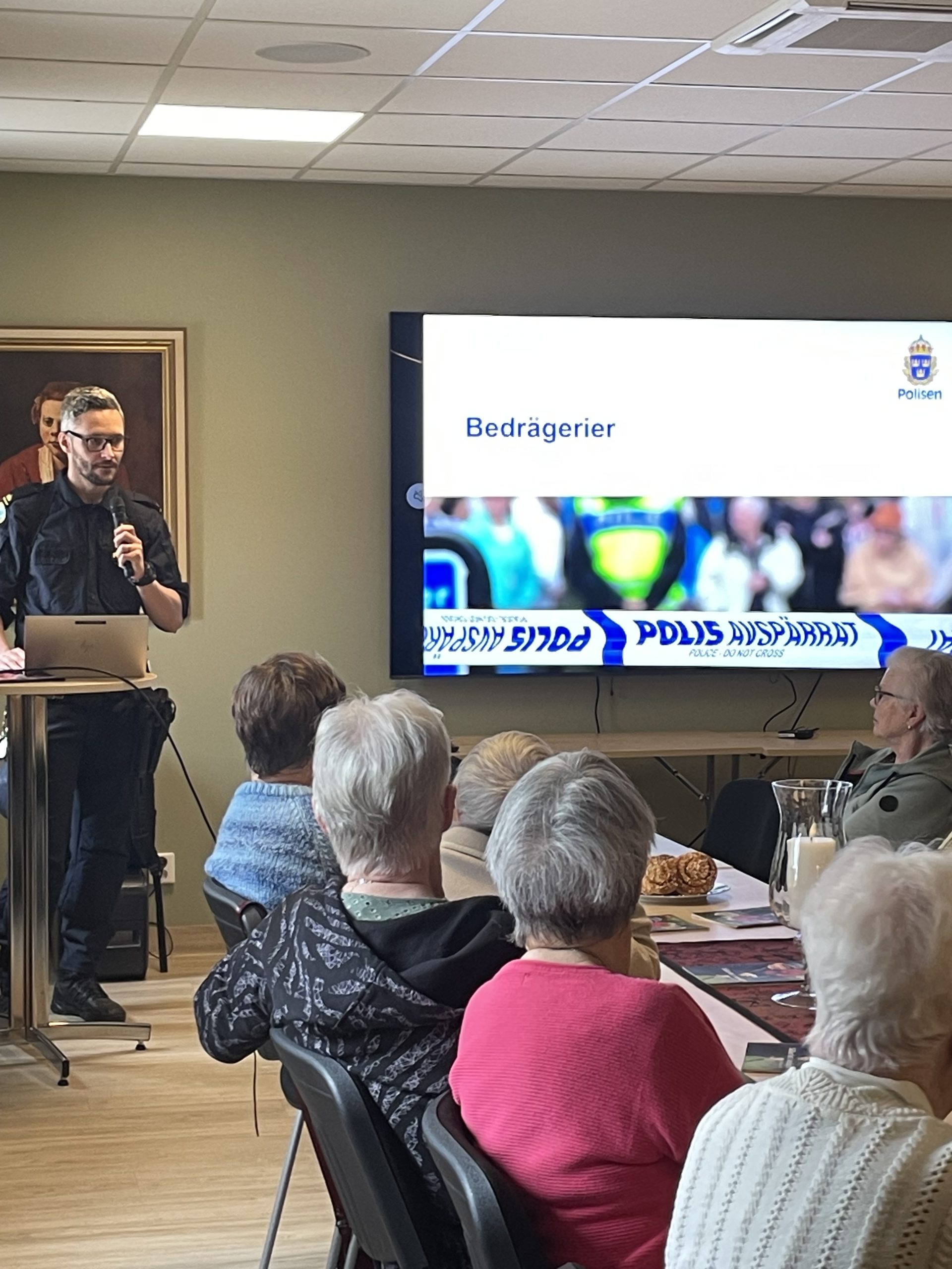 Andreas Lestander från Polisen står bredvid en tv och pratar med seniorer på äldreboendet.